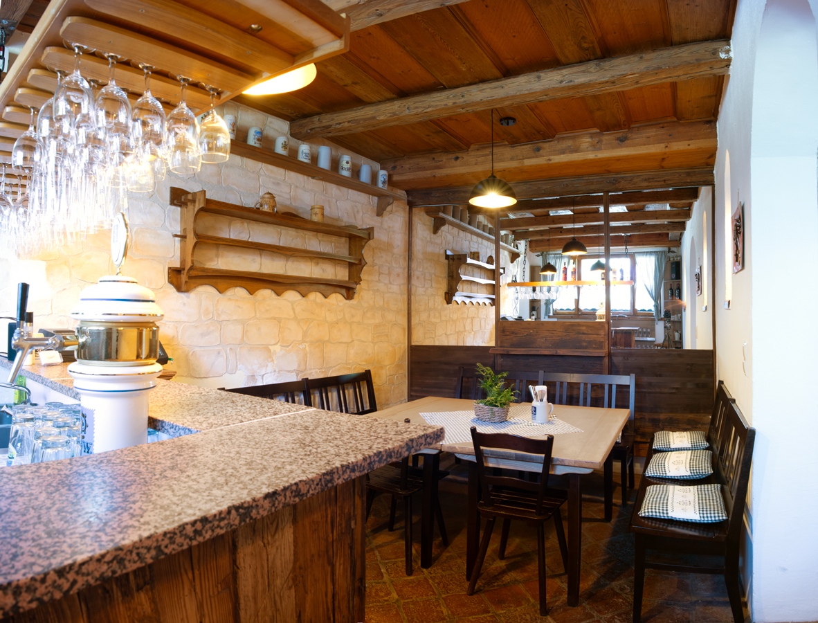 Der Schankraum vom Knödelhof Wirtshaus in Putzbrunn hat eine alte Holzdecke mit Balken, die linke Wand ist mit Natursteinen bestücke. Es gibt nur einen großen Tisch in der Mitte, umgeben mit dunklen Stühlen und Bänken, darauf grün-weiß gewürfelte Kissen. Am Kopfende vom Raum sind Spielge in Holzrahmen integriert, so daß der Raum doppelt so groß wirkt. Links ist die Schank mit Augustiner Bräu Bier. An der Wand hängt ein geschnitztes Holzregal, unter der Decke auf der linken Wand stehen Biersteinkrüge.